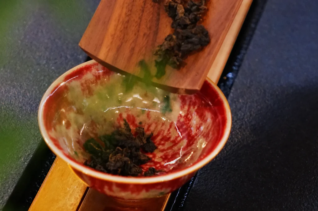 A vibrant red and green ceramic bowl, positioned on a wooden tray, receives loose tea leaves from a wooden scoop. The scene evokes a sense of calm and tradition.