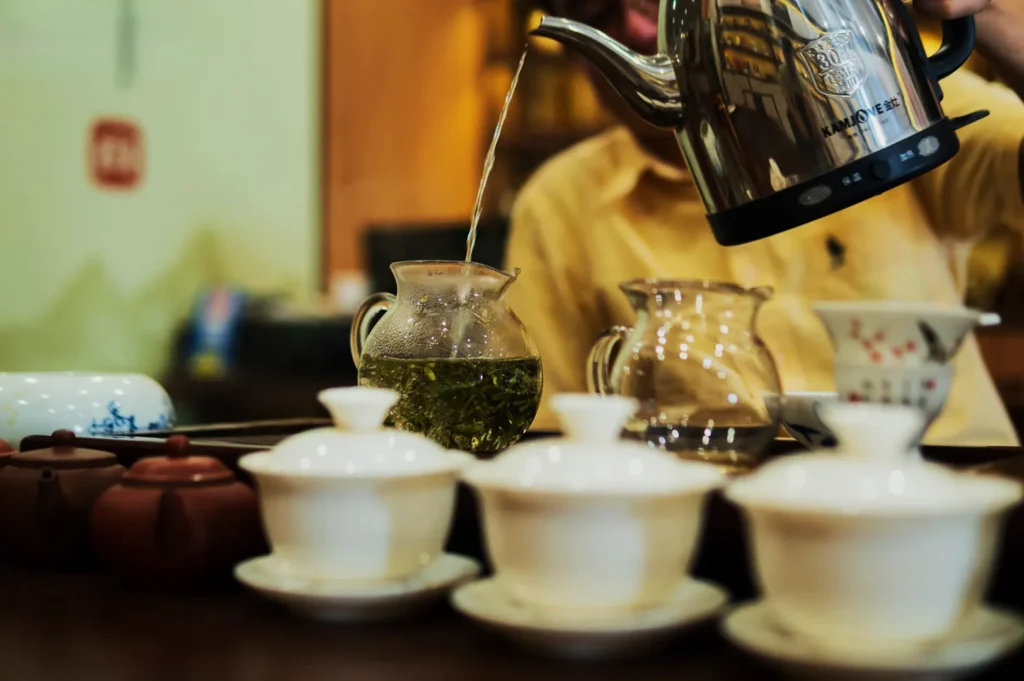 A person pours hot water from a kettle into a glass teapot with green tea leaves. White teacups and clay teapots are arranged on a wooden table.