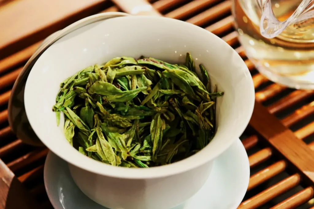 A white bowl filled with vibrant green tea leaves sits on a saucer. Nearby, a glass of light tea rests on a wooden slat table, creating a calming and fresh atmosphere.