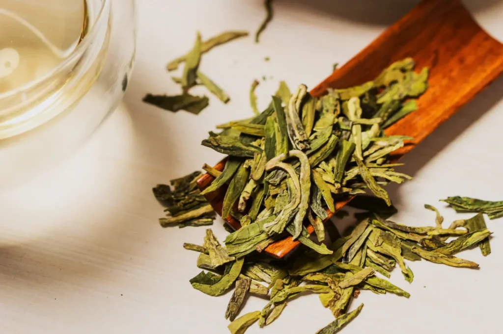 Loose green tea leaves on a wooden scoop next to a glass of tea, on a light surface. The scene suggests tranquility and freshness.