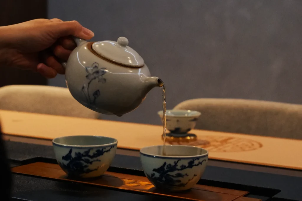 A hand pours tea from a white teapot with floral designs into blue and white teacups on a tray, set in a peaceful, minimalist setting.