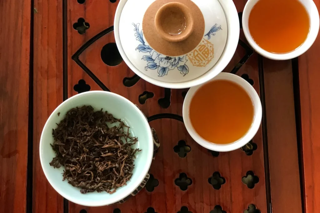 Tea ceremony setup on a wooden table with two cups of amber tea, a gaiwan with a floral design, and a bowl of loose tea leaves, creating a serene atmosphere.