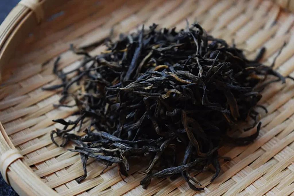 A heap of loose black tea leaves rests on a woven bamboo tray, showcasing their dark, wiry texture. The scene conveys an earthy and natural tone.
