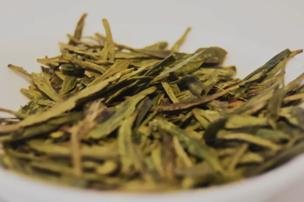 A close-up of dried green tea leaves in a white bowl. The slender, twisted leaves are a vibrant green, conveying freshness and a sense of tranquility.