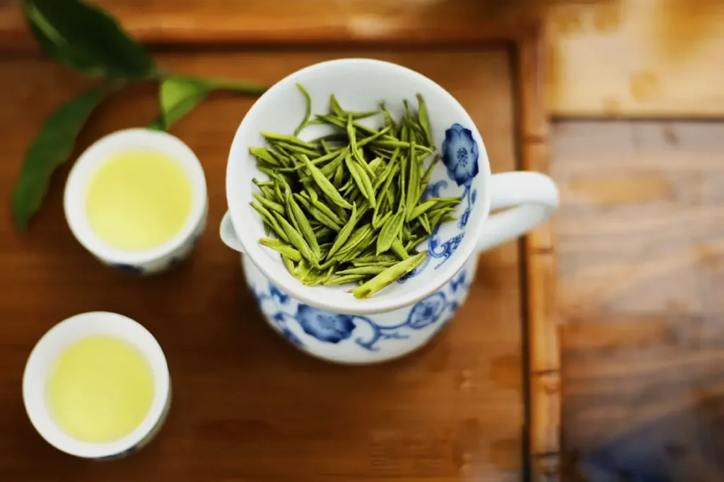 Porcelain cup with floral design holds fresh green tea leaves, placed on a wooden tray. Two small cups of light green tea sit beside it, conveying a calming tone.