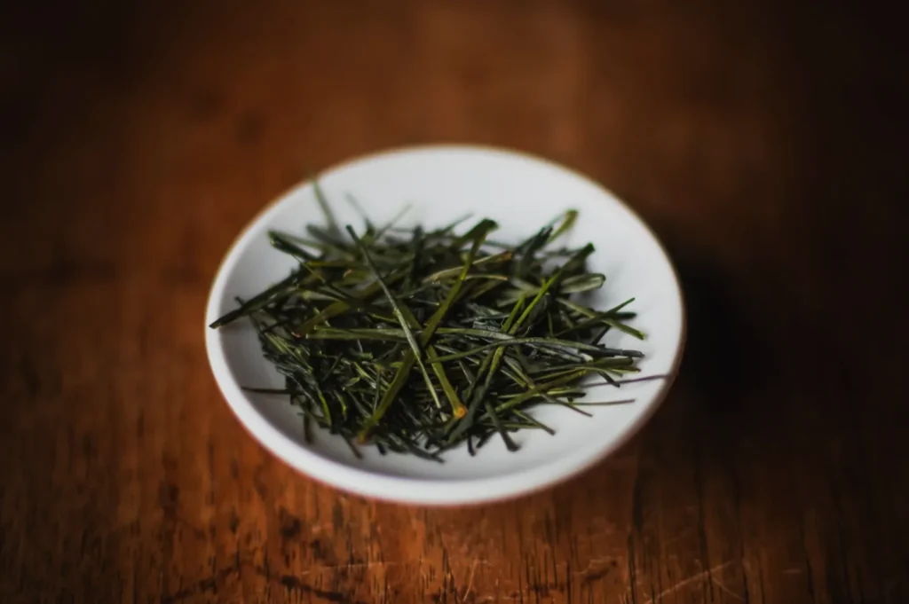 A small white plate on a wooden table holds green tea leaves arranged delicately. The scene conveys a rustic, cozy ambiance.