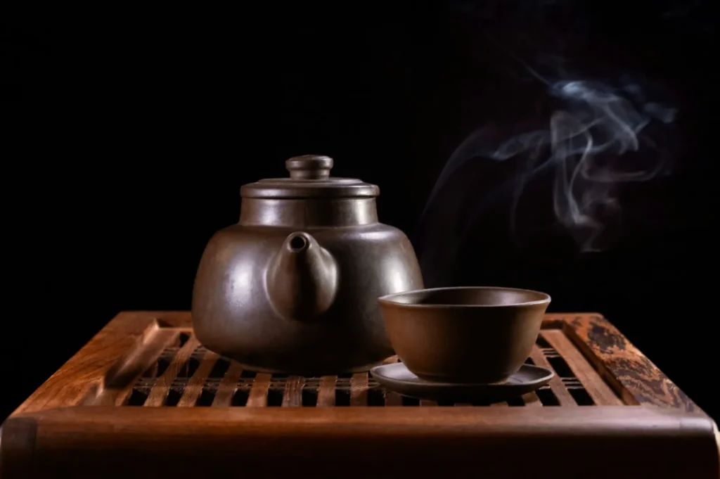 A dark clay teapot and cup set on a wooden tray with wisps of steam rising, against a black background. The scene feels serene and inviting.
