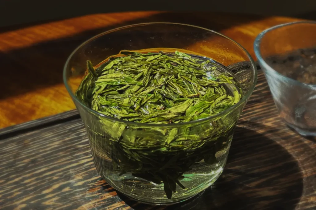 A clear glass filled with vibrant green tea leaves steeping in water, casting a shadow on a wooden surface. Sunlight highlights the fresh leaves.