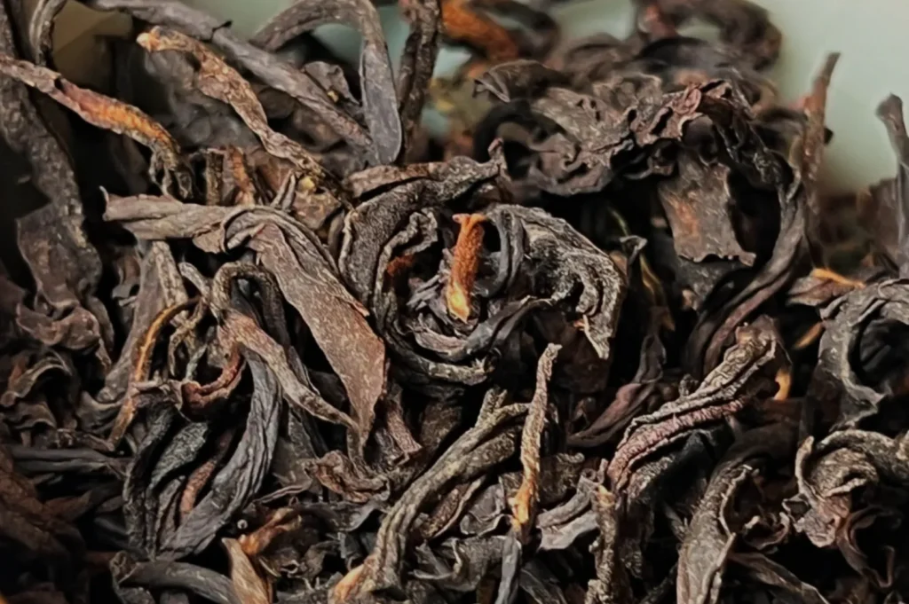 Close-up of curled, dark brown tea leaves, showcasing textured, wrinkled surfaces. The earthy tones suggest a sense of richness and aroma.