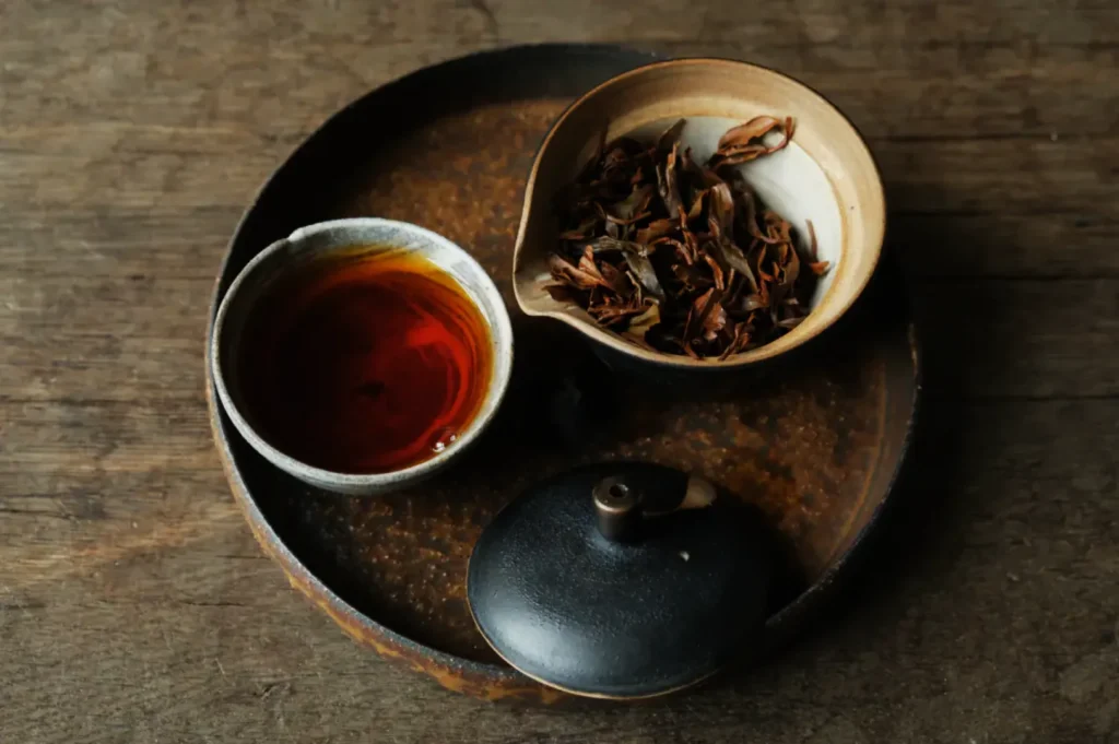 A rustic tea set on a wooden table features a bowl of brewed tea leaves, a small cup of amber tea, and a black teapot lid on a round tray.