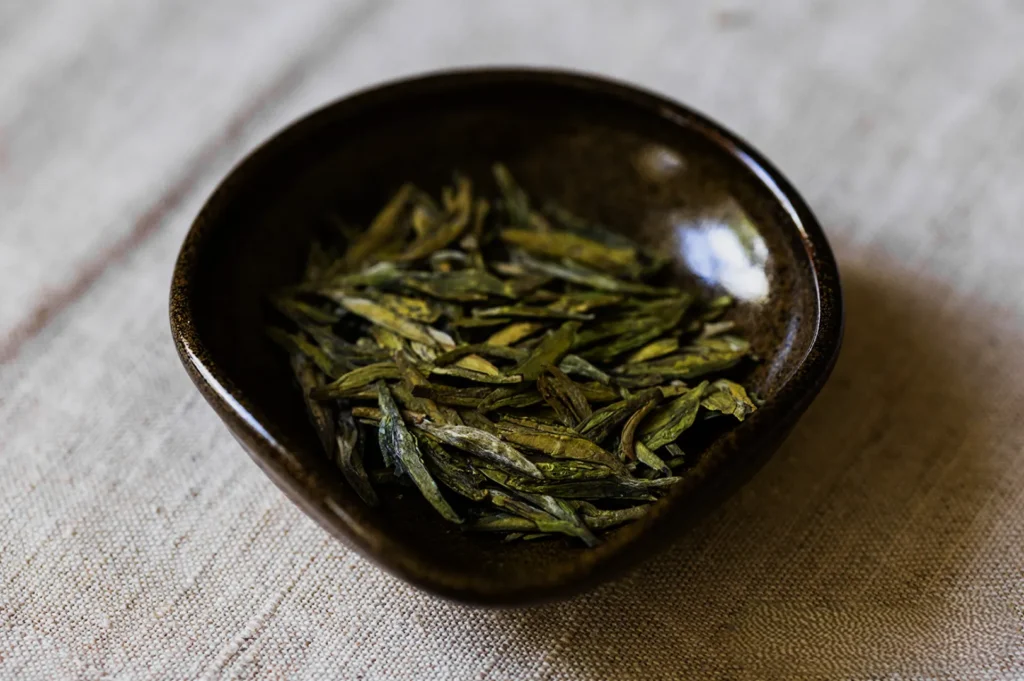 A small brown ceramic bowl holds loose green tea leaves on a textured beige cloth. The setting conveys a calm, natural, and rustic atmosphere.