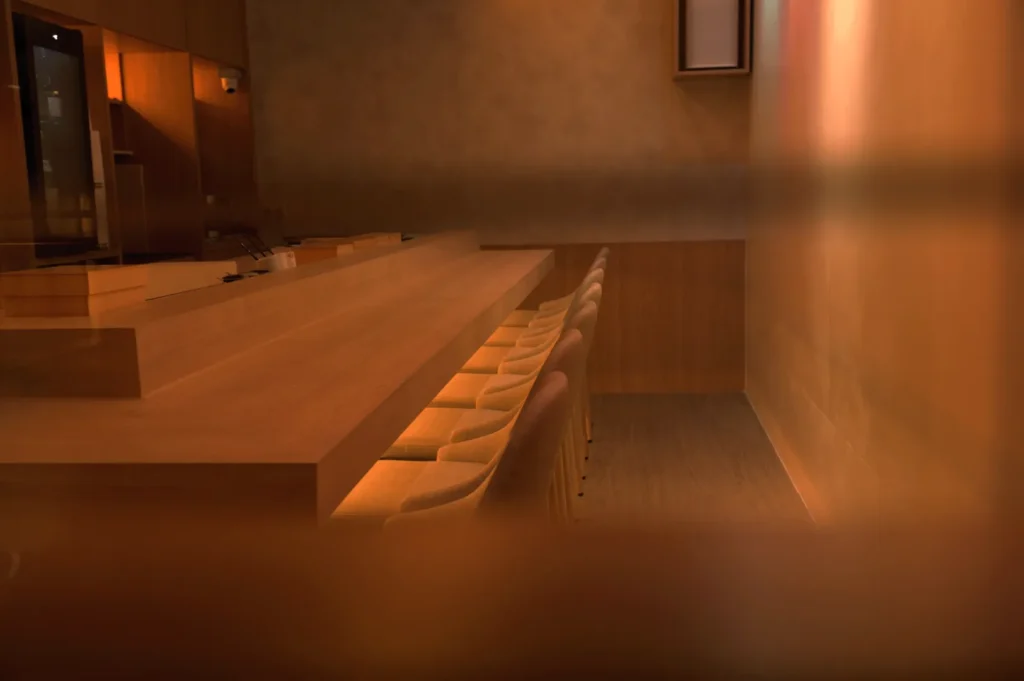 Dimly lit minimalist room with a long wooden counter, lined with cushioned chairs. The warm lighting and wood accents create a cozy, inviting atmosphere.