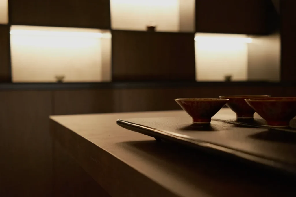 Dimly lit room with three small cups on a tray in the foreground. Shelving with warm backlighting in the background creates a serene ambiance.