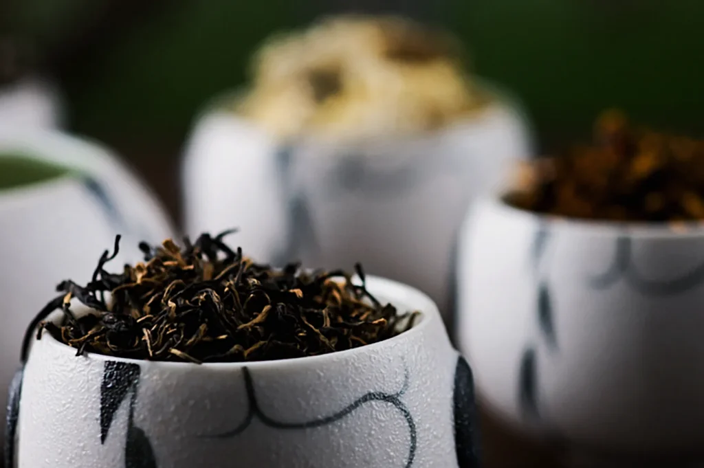 Close-up of a white cup with intricate patterns, filled with dried black tea leaves. Soft focus on other similar cups in the background, creating a calm, inviting atmosphere.