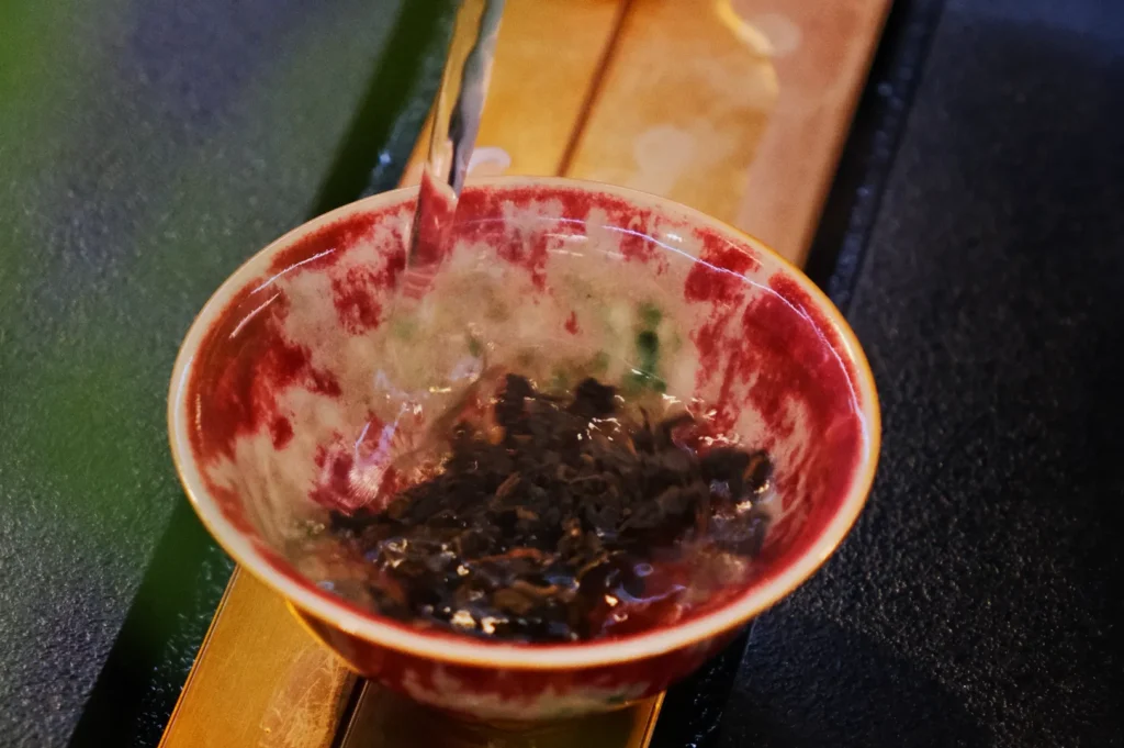 Pouring hot water over loose tea leaves in a red and white ceramic bowl, creating a swirling motion, on a wooden and dark textured surface.