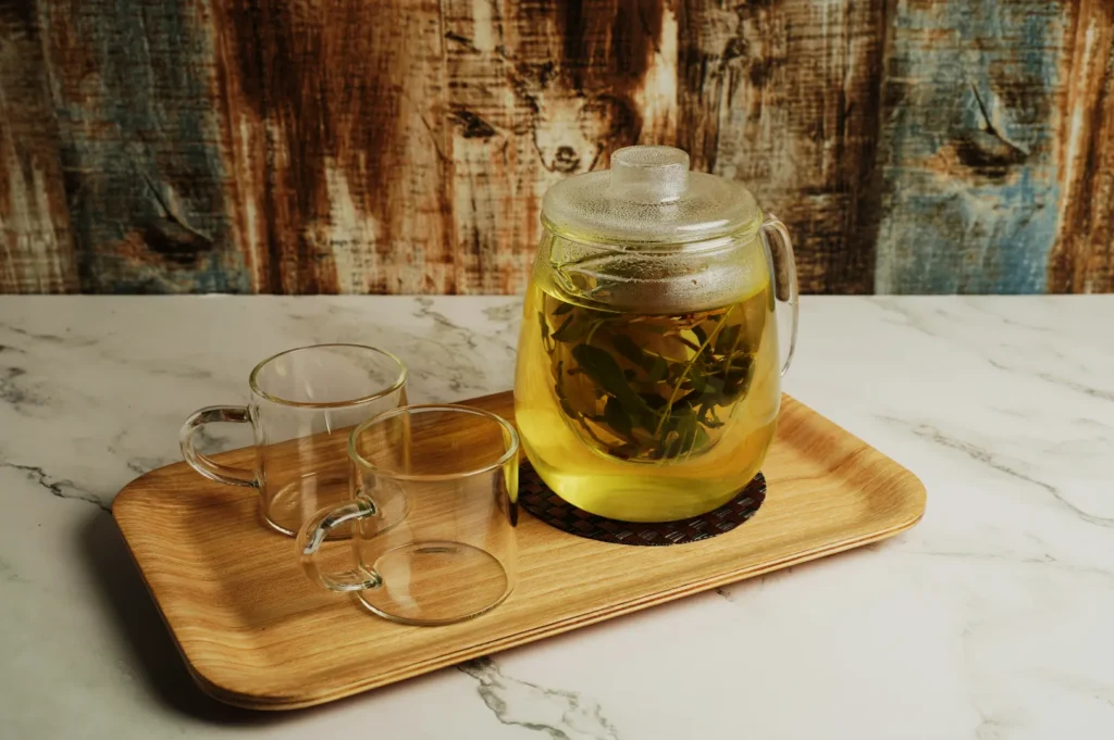 A glass teapot with herbal tea and two empty glass cups sit on a wooden tray. The background features a rustic wood texture, creating a cozy, warm ambiance.