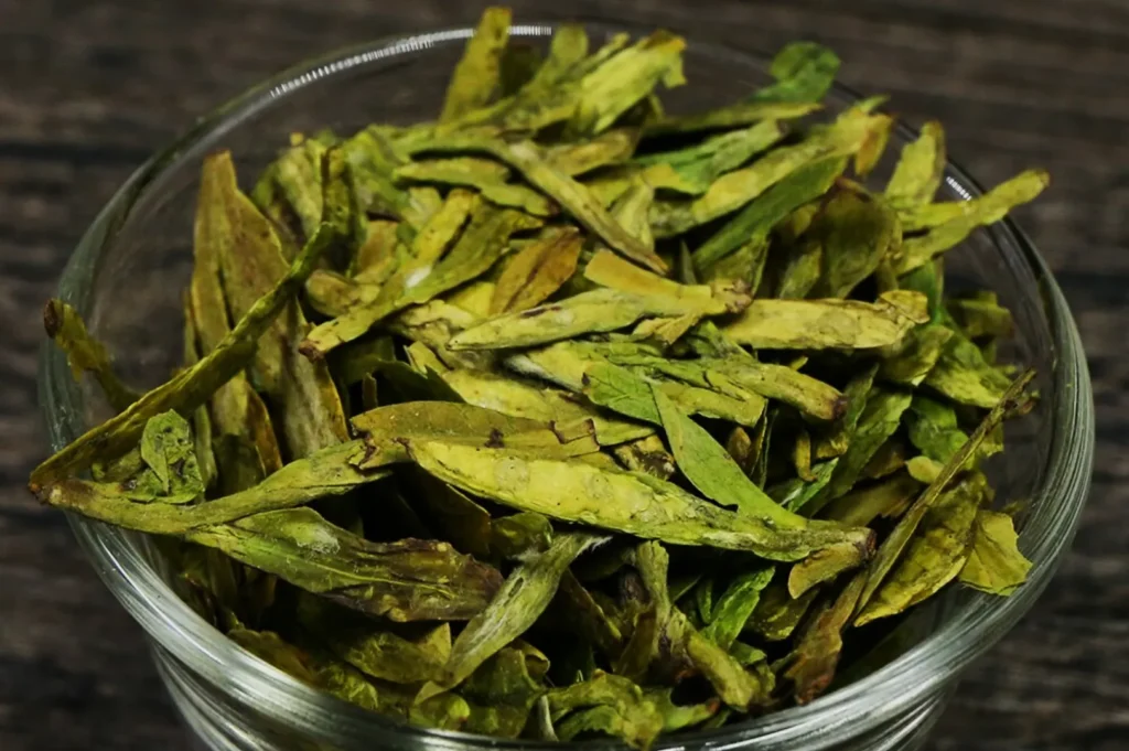 A clear glass bowl filled with dried green tea leaves on a rustic wooden surface. The leaves are elongated and a vibrant green shade, suggesting freshness.