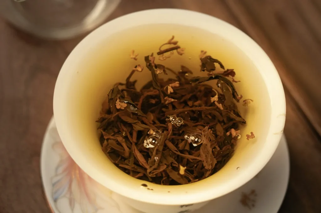 A close-up of brewed tea leaves with small bubbles floating on the surface, set inside a light-colored cup. The mood is warm and calming.