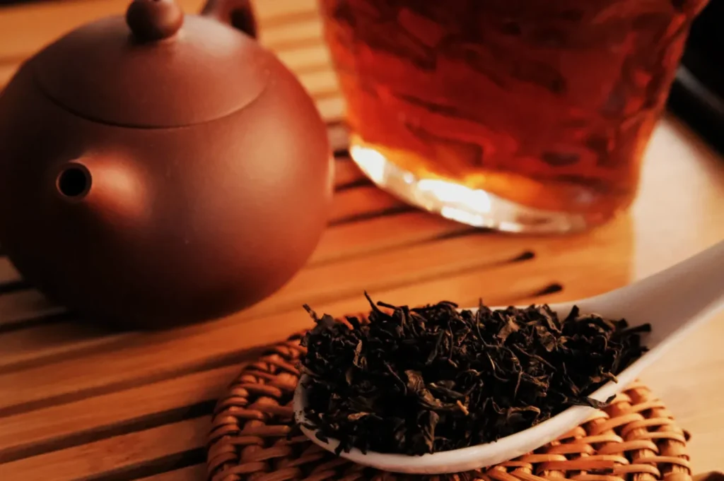 A close-up of a brown teapot, a spoonful of loose black tea leaves, and a glass of tea on a bamboo mat, conveying a warm, cozy atmosphere.