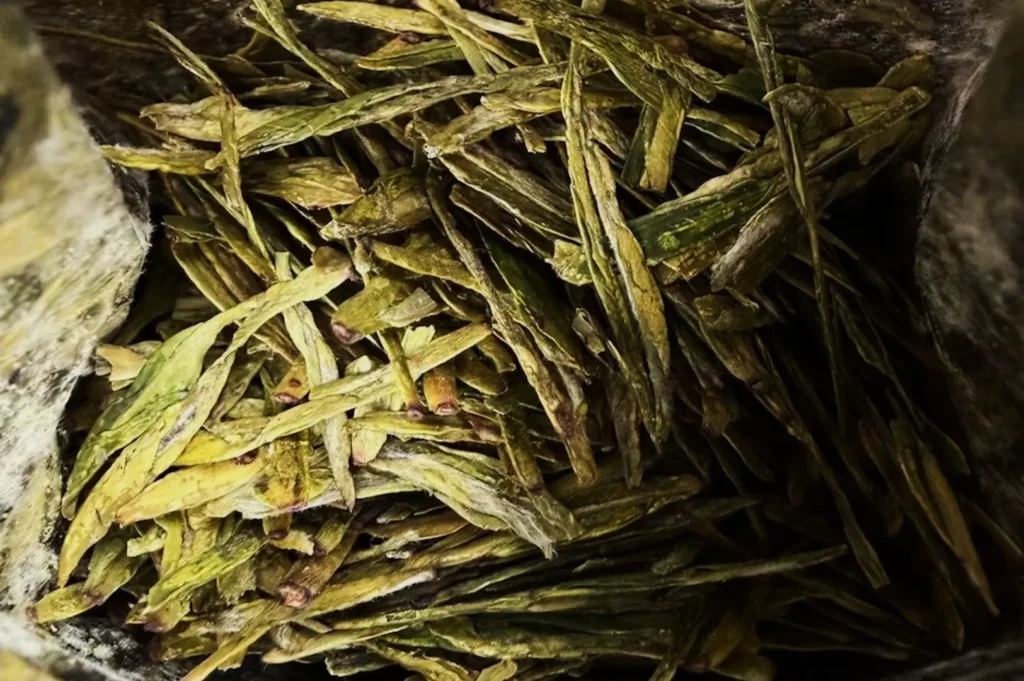 Close-up of dried, flat green tea leaves, scattered in an open bag. The leaves range in shades of green and yellow, creating a textured pattern.