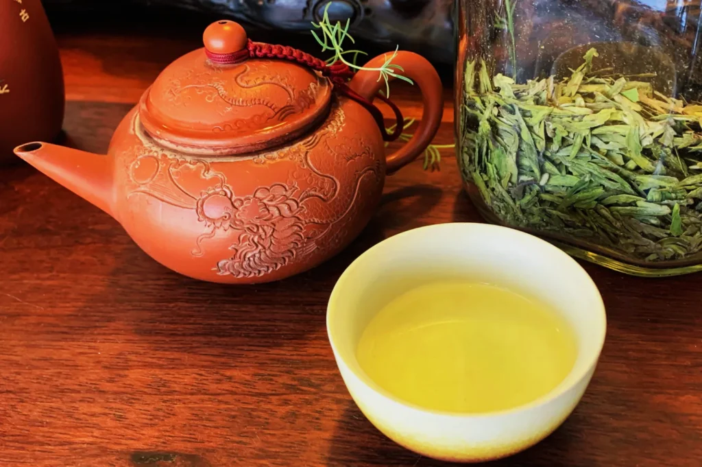 An ornate clay teapot with dragon designs sits beside a glass jar of loose green tea leaves and a bowl of steeped tea on a wooden surface.