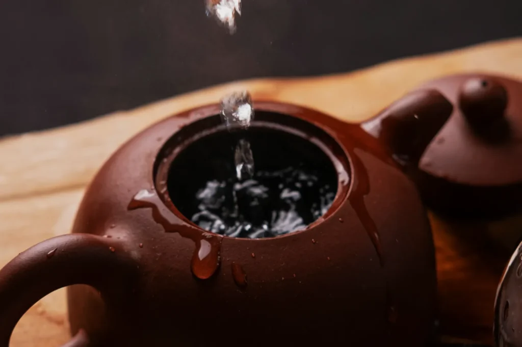 Water is being poured into a clay teapot on a wooden surface, creating ripples. The brown teapot has visible water droplets, suggesting a peaceful, cozy setting.