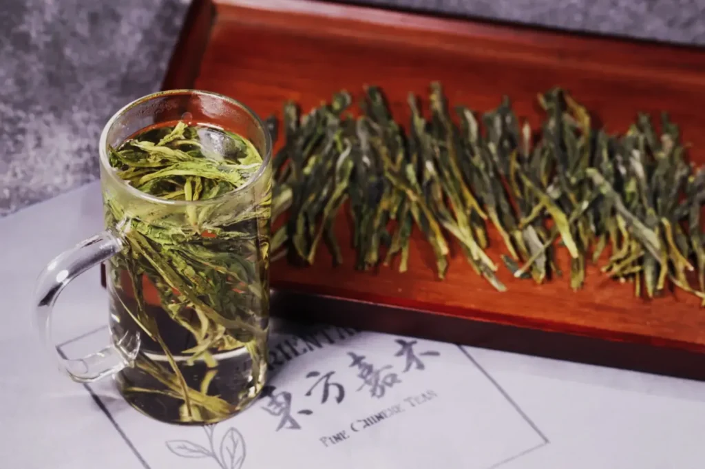 A glass mug filled with clear Chinese tea, showcasing leafy herbs, sits beside a wooden tray with dried tea leaves. The setting is calm and inviting.