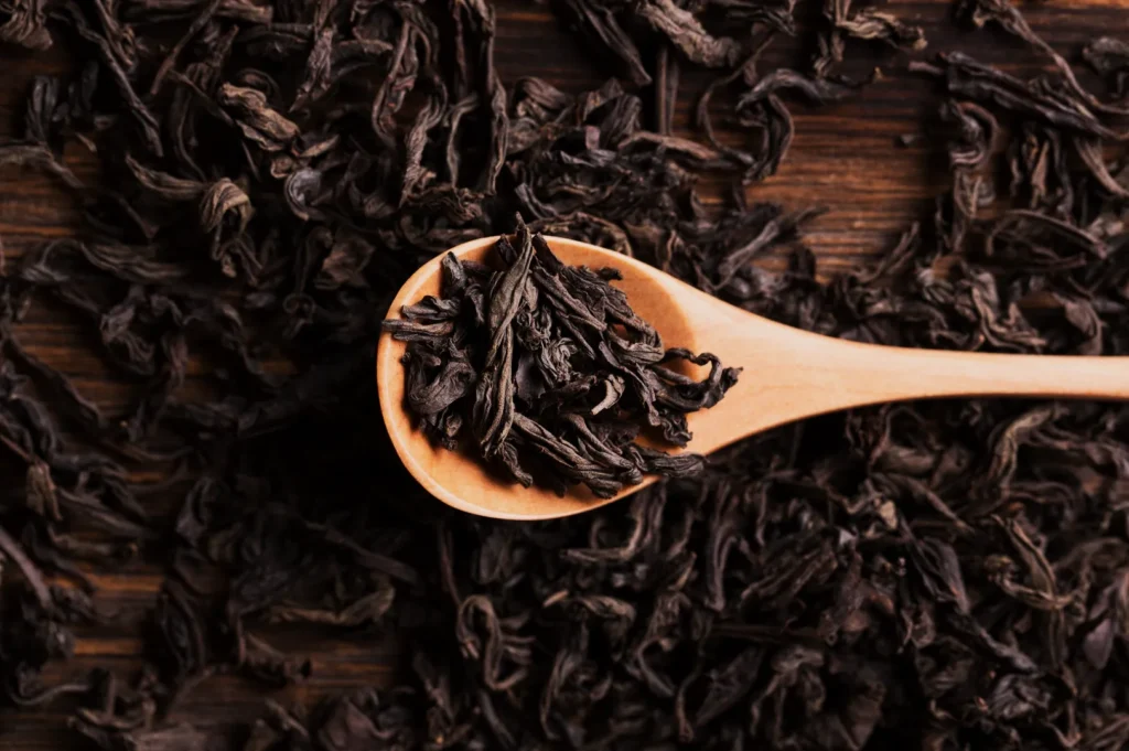 A wooden spoon filled with loose, dark oolong tea leaves rests on a wooden surface, surrounded by more tea leaves, conveying a rustic, earthy feel.