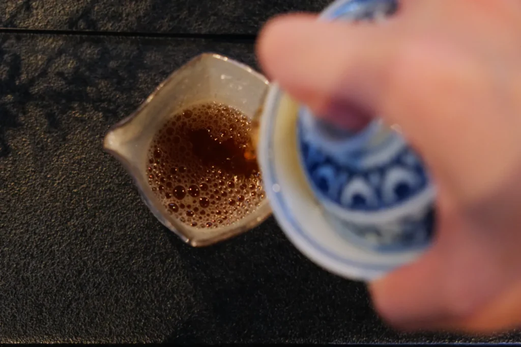 Overhead view of a hand holding a blue patterned cup with a lid, pouring tea into a cream-colored pitcher on a dark textured surface.