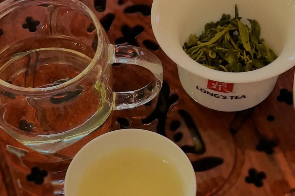 A clear glass teapot with water, a bowl of steeped green tea leaves marked "Long's Tea," and a small cup of light yellow tea on a wooden table.
