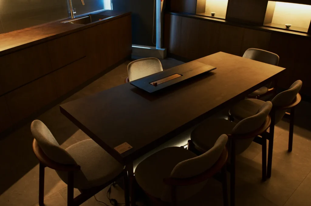 Modern dining area with six grey cushioned chairs around a rectangular wooden table. Soft ambient lighting creates a warm, cozy atmosphere.