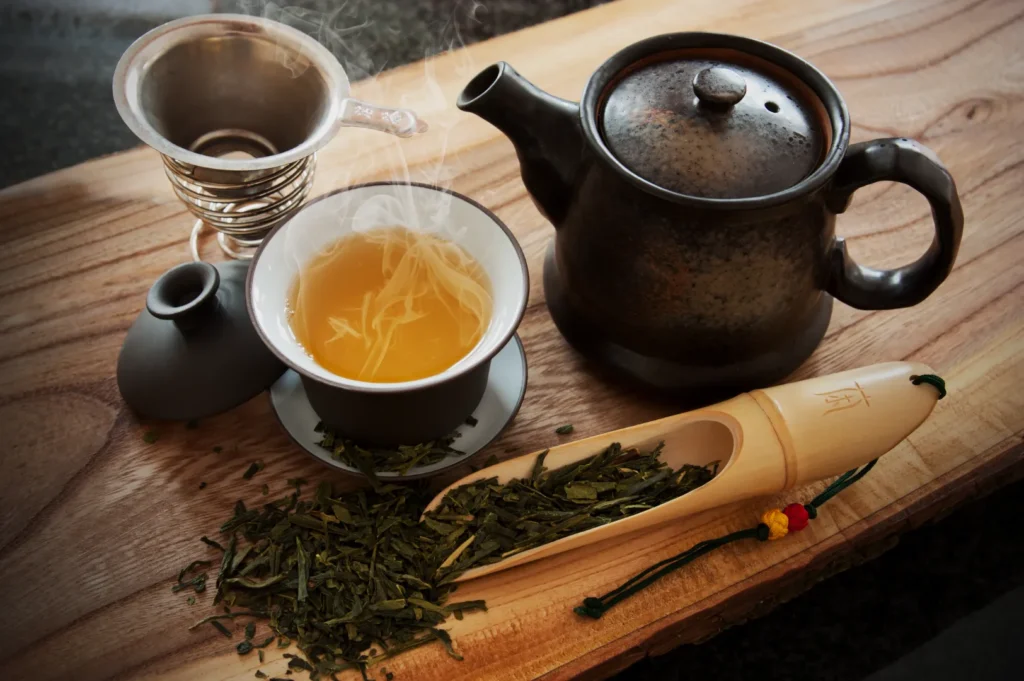 Steaming cup of green tea on a wooden board with a black teapot, tea strainer, and bamboo scoop holding loose leaves. Cozy and inviting atmosphere.