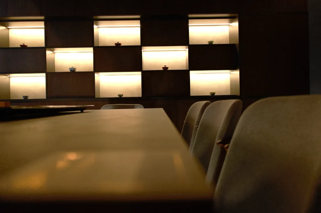 Dimly lit room with a modern, minimalist design. Illuminated shelves display small decorative items. Soft chairs line a dark wooden table, creating a cozy atmosphere.