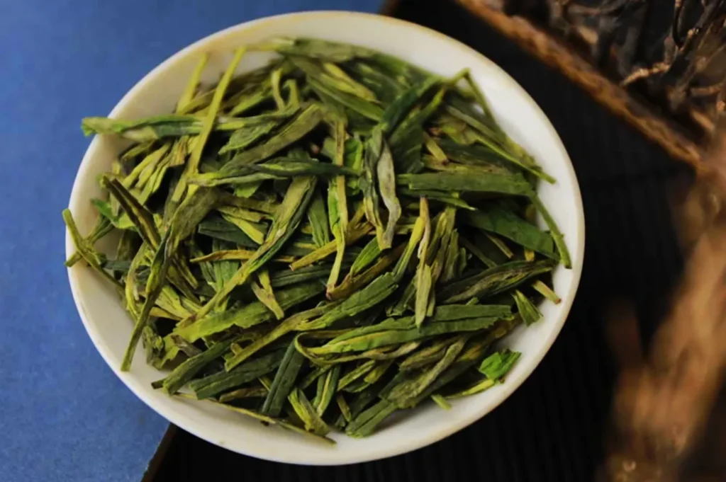 A white bowl filled with long, flat green tea leaves sits on a dark surface. The vibrant green tones convey freshness and aroma, suggesting quality tea.