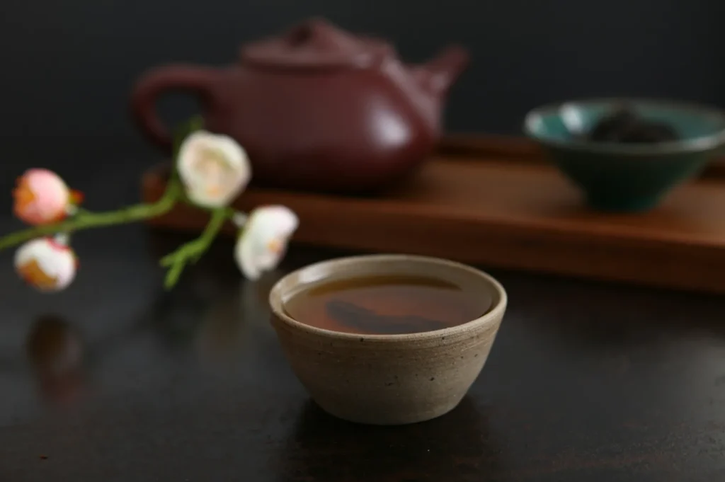 A ceramic teacup filled with tea sits on a dark surface. Behind it, a clay teapot and a green bowl on a wooden tray are blurred. Soft lighting creates a calm ambiance.