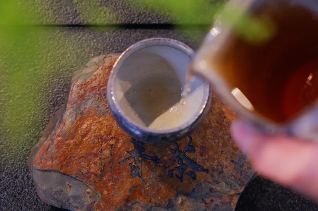 Pouring tea into a ceramic cup atop a textured stone coaster with engraved calligraphy, surrounded by a calming, soft-focus garden setting.