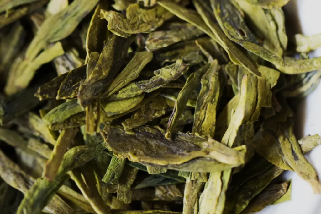 Close-up of dried tea leaves, primarily green with slight hues of brown, layered in a random pile. The texture appears crisp and papery.