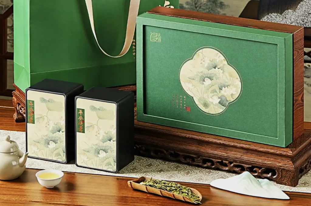Elegant tea set display on a wooden tray: green gift box with floral design, matching tins, white teapot, cup, and loose tea leaves. Calm and refined tone.