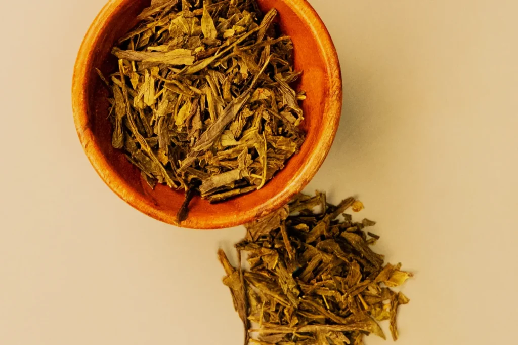 A small wooden bowl filled with loose green tea leaves on a light background. Some tea leaves are scattered beside the bowl, creating an earthy, natural feel.