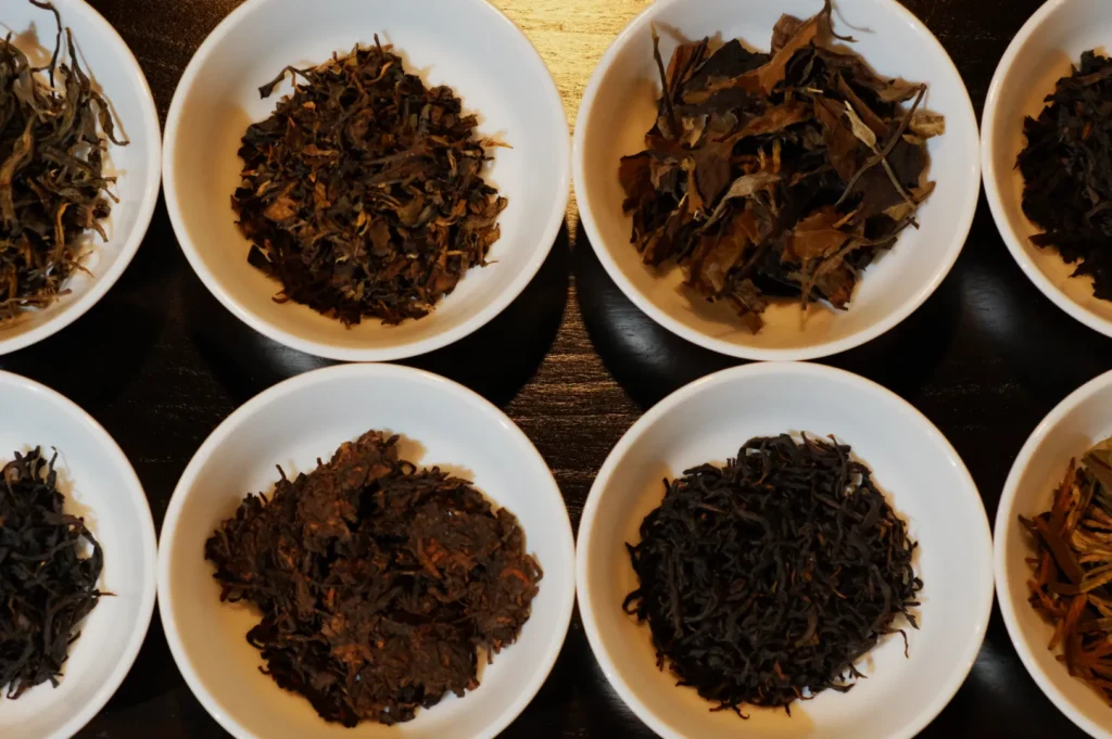 Eight white bowls arranged in two rows on a dark surface, each filled with different varieties of dry, loose tea leaves, showcasing diverse textures and shades.