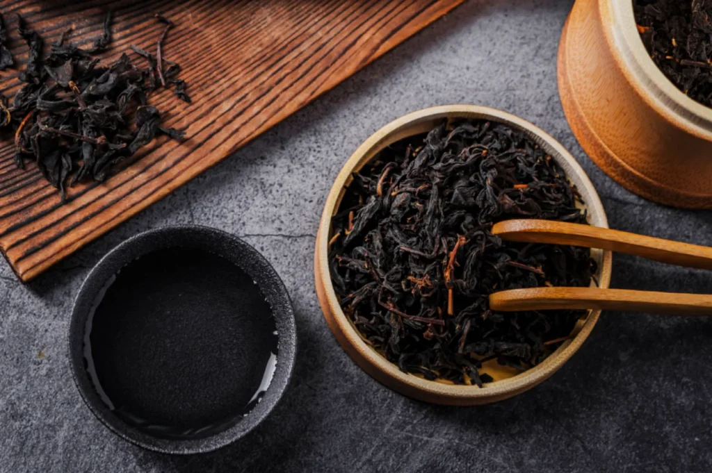 A wooden bowl of dried tea leaves with chopsticks, a black cup of water, and more tea leaves on a wooden board, all on a gray textured surface. Relaxing and earthy tone.