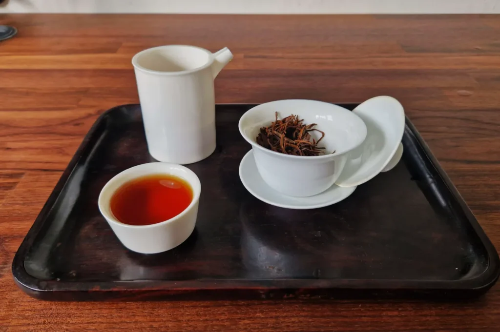 A dark wooden tray holds white ceramic teaware: a cup with brewed tea, a small pitcher, and a bowl with dry tea leaves, set on a wooden table.