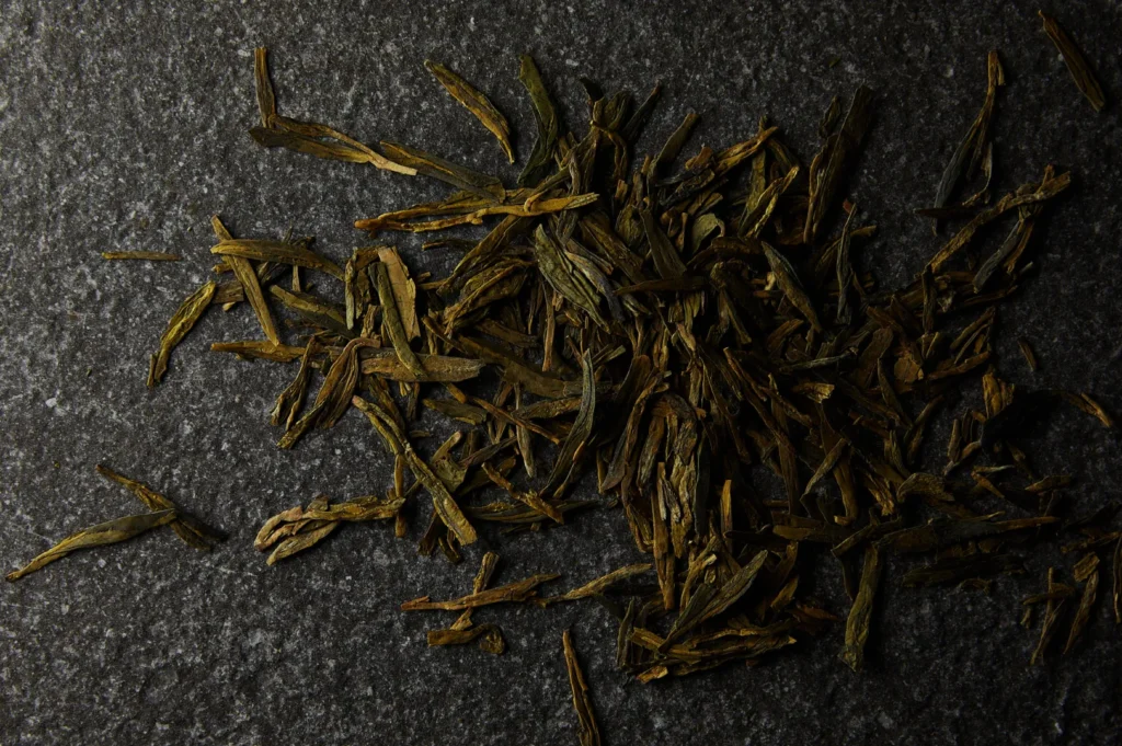Dried green tea leaves scattered on a dark, textured surface. The leaves are long and flat, creating an earthy, natural feel.