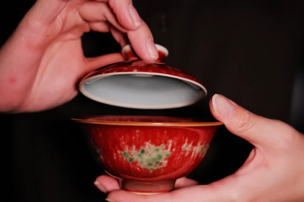 Hands gently lift the lid of a red, ornate porcelain tea bowl, revealing its interior. The scene conveys care and craftsmanship.