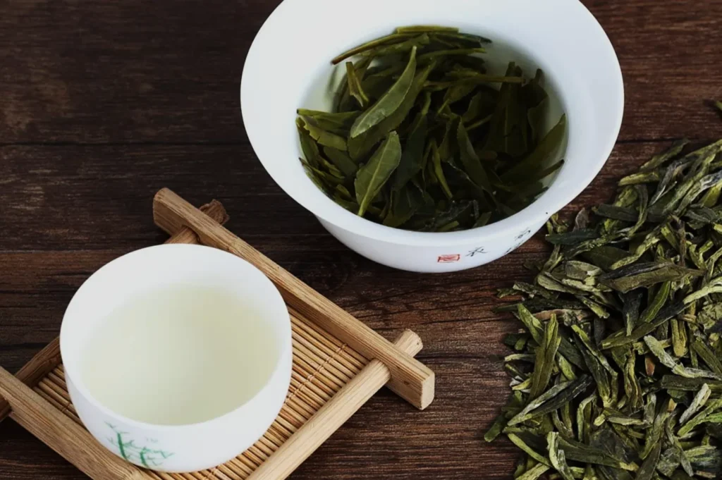 A white teacup with light green tea sits on a bamboo mat beside a bowl of steeped green tea leaves. Loose tea leaves scattered on a wooden table.