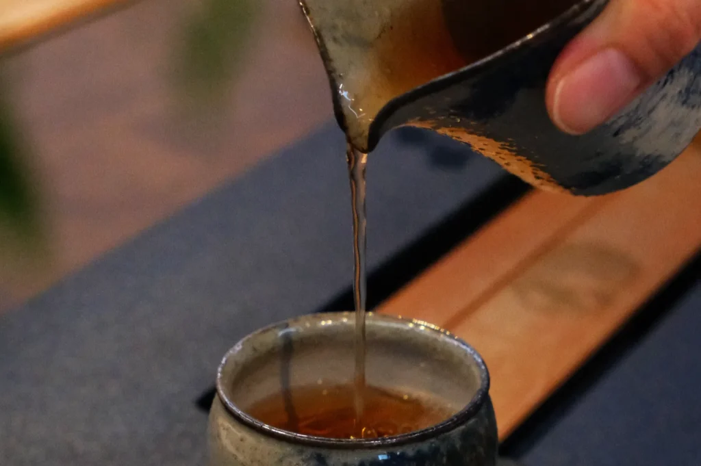 A close-up of tea being poured from a small, rustic ceramic pitcher into a matching cup. The scene is calm, with natural tones and textures.