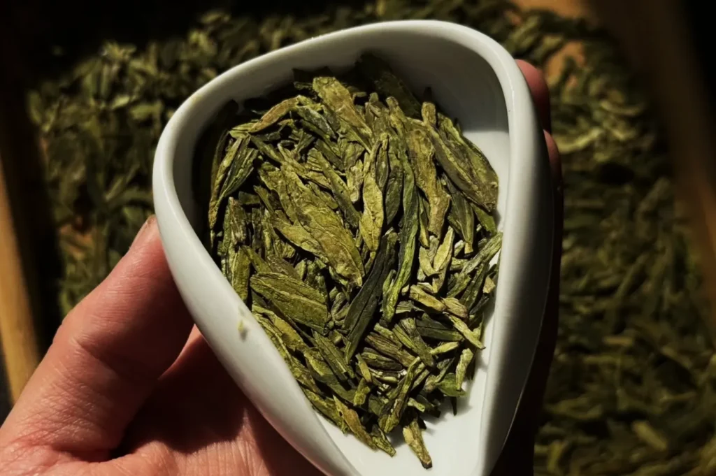 A hand holds a white, heart-shaped bowl filled with green tea leaves. Sunlight highlights the leaves' texture, casting shadows. Earthy, warm tone.
