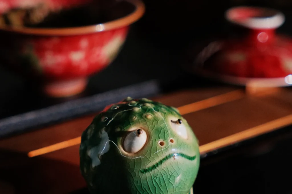 A small, green ceramic frog with an expressive face sits on a wooden surface, surrounded by red, traditional-style bowls. The scene conveys a whimsical mood.