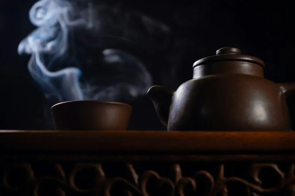 A clay teapot and cup sit on a carved wooden surface, with ethereal smoke swirling against a dark background, creating a tranquil, aromatic atmosphere.