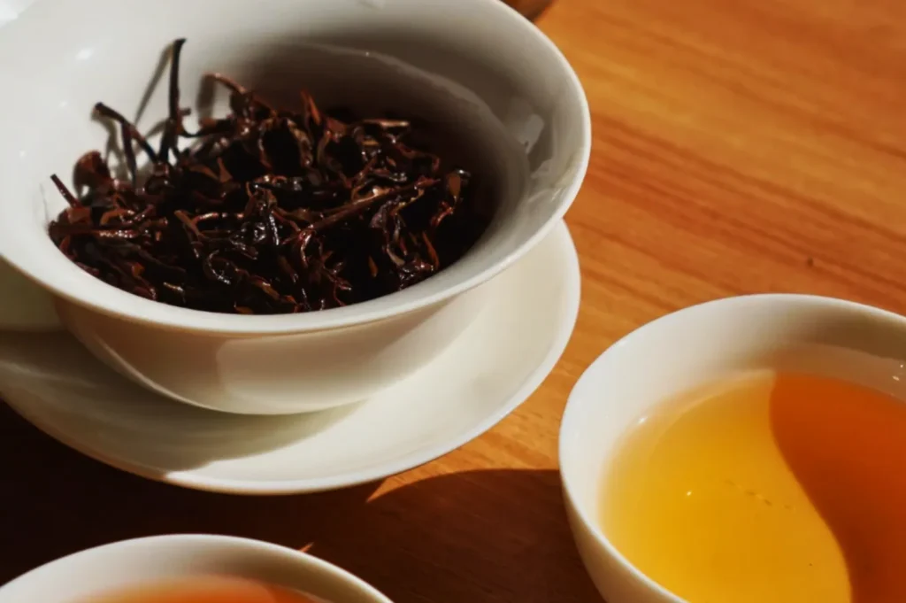A white bowl with dried black tea leaves sits on a wooden table beside a white cup filled with warm amber tea, creating a cozy and relaxed atmosphere.